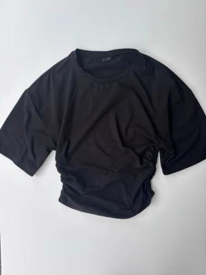 Svart t-shirt med scrunch i sidan - En svart t-shirt med korta ärmar och rund hals. Tillverkad i mjukt bomullsmaterial för skön känsla och avslappnad passform. Knappt använd 
