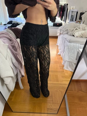 Svarta spetsleggings med raka ben - Säljer ett par svarta leggings i genombruten spets med hög midja och raka ben. De har elastisk midja och är helt genomskinliga, inbyggda shorts. Materialet är stretchigt och följsamt för en bekväm passform.