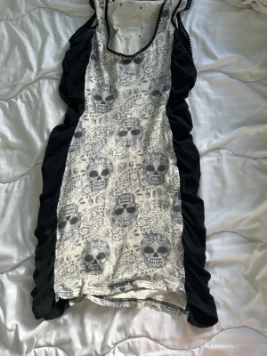 Kort klänning med döskallemönster - Unik kort klänning från Pussy Deluxe med svartvita döskallar och paisleymönster. Klänningen har svarta sidor och en figurnära passform. Rund halsringning med dekorativ kant. Perfekt för dig som gillar alternativ stil!
