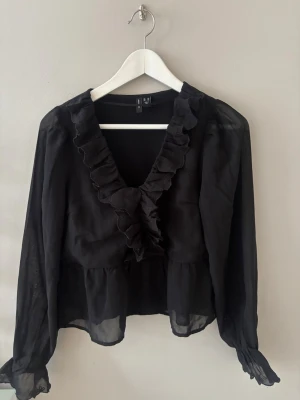 Svart volangblus från Vero Moda - Snygg svart blus från Vero Moda med transparenta långärmar och volangdetaljer längs den djupa V-ringningen. Tillverkad i ett lätt och luftigt material som ger en feminin och trendig look. Perfekt att styla med jeans eller kjol.