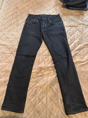 Svarta raka jeans från Nudie Jeans - Säljer ett par svarta jeans från Nudie Jeans med rak passform och klassisk femficksdesign. Jeansen har silverfärgade nitar och är tillverkade i ett slitstarkt denimtyg. Perfekta för dig som gillar en clean och tidlös look.