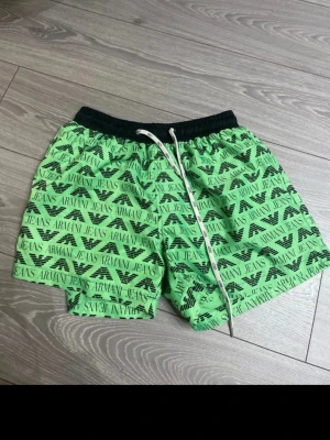 Gröna badbyxor från Armani Jeans - Snygga gröna badbyxor från Armani Jeans med svart resårmidja och vit dragsko. Badbyxorna har ett all-over tryck med logga och text samt meshfoder på insidan för extra komfort. Perfekta för stranden eller poolen.