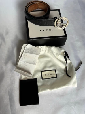 Äkta Svart Gucci skärp - Gucci skärp för man i svart läder med präglad logotyp och rund, silverfärgad GG-spänne. Skärpet levereras med tygpåse och tillhörande kartong samt ett kvitto från när den var köpt i deras butik i Italien. Bältet har storleken på 85•34.