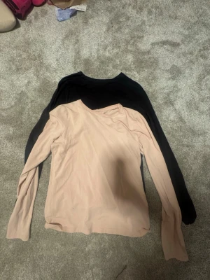 Två långärmade toppar från H&M - Säljer två basic långärmade toppar från H&M, en i ljus beige och en i svart. Båda har rund halsringning och är tillverkade i mjuk bomull, perfekta att styla till jeans eller kjol. Enkla och stilrena plagg för alla tillfällen. Storlek 170 passar som S-M
