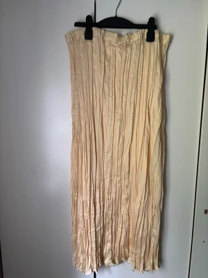 Plisserad beige långkjol från H&M - Säljer en plisserad beige långkjol från H&M. Kjolen har resår i midjan och är tillverkad i ett lätt, glansigt material som ger ett snyggt fall. Perfekt för dig som gillar en stilren och trendig look.