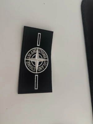 Stone Island badge - white - Säljer ett svart tygmärke från Stone Island med den klassiska vita kompassen broderad i mitten. Märket är rektangulärt och har vita detaljer och text. Perfekt att sy eller fästa på jackor, väskor eller andra plagg.