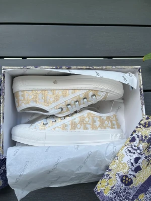Beige och vita Dior sneakers - Snygga sneakers från Christian Dior i beige och vitt med det klassiska Dior-mönstret på sidan. Skorna har vita snören, vit sula och detaljer med logotypen på sidan och tungan. Tillverkade i textil och gummi för en trendig och exklusiv look.