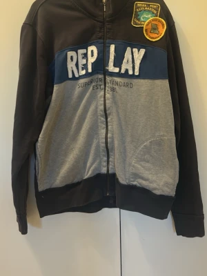 Replay hoodie med patchar och dragkedja - Snygg hoodie från Replay i svart, grått och blått med dragkedja framtill. Stor REPLAY-text i vitt på bröstet och coola patchar på axeln. Tillverkad i mjuk bomullsblandning och har fickor framtill. Perfekt för en avslappnad streetwear-look.