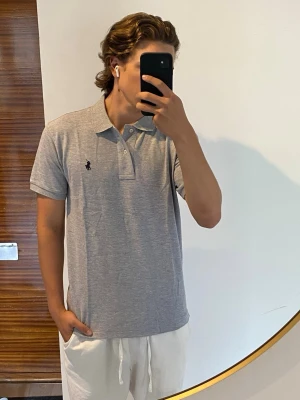 pikétröja från Ralph Lauren - Klassisk grå pikétröja från Ralph Lauren med den ikoniska broderade loggan på bröstet. Tröjan har korta ärmar, krage och knappar framtill. Tillverkad i mjuk bomull för en bekväm känsla och stilren look. Storlek M, jag är 176 cm.