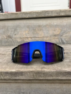 Blå sportiga solglasögon - Säljer ett par riktigt snygga sportiga solglasögon med spegelblått, kantigt glas och svart båge. De har en futuristisk vibe och passar perfekt för dig som vill sticka ut. Linsen är bred och ger bra täckning för ögonen.