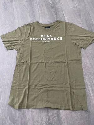 Olivgrön Peak Performance t-shirt - Snygg olivgrön t-shirt från Peak Performance med vit logga tryckt på bröstet. Klassisk rund hals och korta ärmar. Tillverkad i mjuk bomull som känns skön mot huden. Perfekt till jeans eller shorts för en avslappnad look.