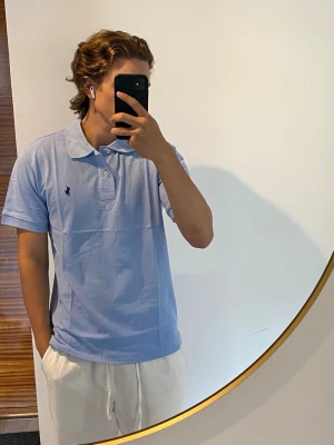  pikétröja från Ralph Lauren - Säljer en ljusblå pikétröja från Ralph Lauren med klassisk krage och två knappar framtill. Tröjan har den ikoniska mörkblå loggan broderad på bröstet och är tillverkad i mjuk bomull. Perfekt till jeans eller shorts för en clean look. Storlek m, är jag 176 cm