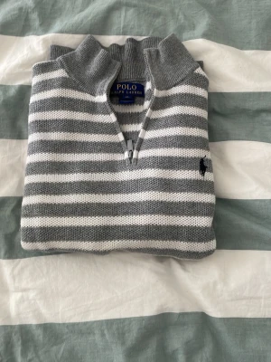 Gråvit randig stickad tröja från polo - Snygg stickad tröja från Polo Ralph Lauren i grått och vitt med horisontella ränder. Tröjan har halv dragkedja, hög krage och den klassiska broderade loggan på bröstet. Perfekt för dig som gillar stilren och tidlös design!😇😎passar dig som är runt 155-160 cm 45kg 