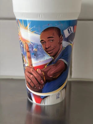 Färgglad plastmugg med fotbollstema - Säljer en plastmugg med färgstarkt tryck av tecknade fotbollsspelare och texten 'HENRY' i stora bokstäver. Motivet har blå, gul, röd och vit färg och visar en spelare i aktion samt publik i bakgrunden. Perfekt för dig som gillar sportiga och unika muggar.