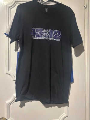 Svart t-shirt med tryck 1312 - Svart t-shirt med stort lila och vitt tryck på bröstet där det står '1312' och 'Försök aldrig stoppa vårt stöd'. Klassisk rund hals och korta ärmar. Materialet känns som bomull och passformen är normal. Inget lags merch utan egentryckt som ska återvinnas i supporterkulturen. Komemr köpas in fler vid intresse. Finns en inne just nu
