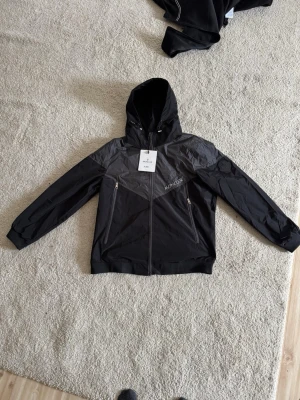 Svart vindjacka från Moncler - Snygg svart vindjacka från Moncler med huva och dragkedja framtill. Jackan har två snedställda fickor med dragkedja och en diskret logotyp på bröstet samt patch på ärmen. Tillverkad i lätt, vindtätt material som passar perfekt till streetwear.                                           Står XL men den är liten i storlek passar S/M skulle jag säga.