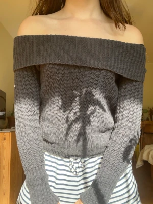 Mörkgrå off shoulder stickad tröja - Trendig mörkgrå stickad tröja med off shoulder-design och bred ribbad kant upptill. Tröjan har lång ärm och är gjord i ett mjukt, grovstickat material som ger en avslappnad men snygg look. Perfekt för dig som vill ha en modern och bekväm stil.