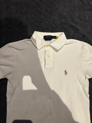 Vit pikétröja från Ralph Lauren - Klassisk vit pikétröja från Ralph Lauren med korta ärmar och krage. Tröjan har två knappar framtill och den ikoniska broderade loggan i beige på bröstet. Tillverkad i bomull för en bekväm och stilren look.