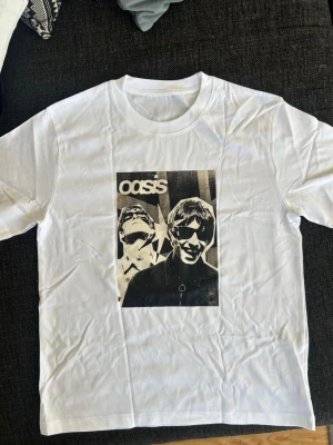 Vit Oasis t-shirt med tryck - Vit t-shirt med svartvitt tryck av bandet Oasis på bröstet. Klassisk rund hals och korta ärmar. Perfekt för dig som gillar musik och vill ha en stilren bandtröja.