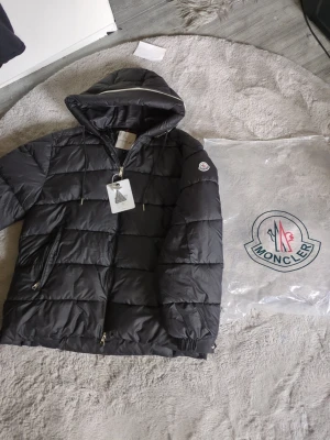Svart dunjacka med huva från Moncler - ( står S men passar helt klart som en xxl+ för dig som väger mellan 110 och 120kg passar den perfekt)Snygg svart dunjacka från Moncler med huva och dragkedja framtill. Jackan har en quiltad design, två sidofickor med dragkedja och Moncler-logga på ärmen. Perfekt för kalla dagar och riktigt varm tack vare dunfyllningen.