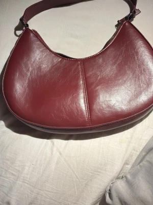 Vinröd axelväska i skinnimitation - Snygg vinröd axelväska i skinnimitation med halvmåneform och bred axelrem. Väskan har en enkel design med synlig söm i mitten och dragkedja upptill. Perfekt för dig som vill ha en stilren och trendig accessoar.
