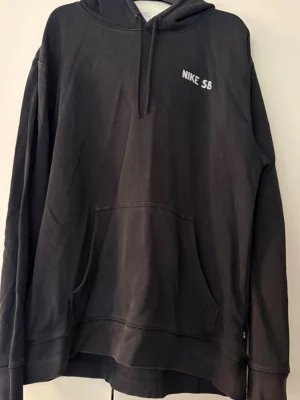 Svart Nike SB hoodie med huva - Svart hoodie från Nike SB med klassisk huva och dragsko. Framtill finns en vit Nike SB-logga på bröstet och en stor känguruficka. Baktill på ryggen finns också tryck/logga. Hoodien har en avslappnad passform och är tillverkad i mjukt material, perfekt för en chill och sportig stil. Svart, storlek M.