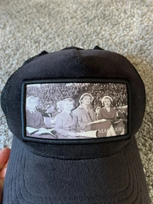 Svart truckerkeps med fototryck - Säljer en svart truckerkeps med mesh på sidorna och ett stort svartvitt fototryck framtill på kungen som ung. Kepsen har böjd skärm och justerbar snapback. Perfekt för dig som gillar streetwear och unika detaljer.