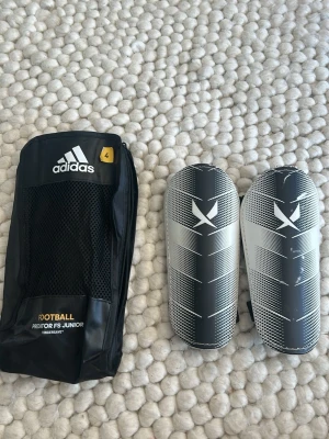 Adidas benskydd junior svart/vit - Säljer ett par Adidas benskydd för juniorer i svart och vitt med grafiskt mönster. Kommer med originalförpackning. Perfekta för fotbollsträning och matcher.