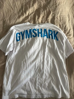 Vit Gymshark t-shirt med blå text - Vit t-shirt från Gymshark med tryck. As snygg att ha på gymmet! Finns matchande shorts/sport bh i min profil om man vill köpa bundle. Den är unisex. Den är true to size men passar flera storlekar beroende på hur man vill ha den. Skriv för frågor/bilder💕