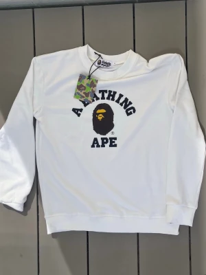 Bape Tröja - Vit sweatshirt med svart A Bathing Ape-tryck och ikoniskt apansikte i svart och gult på bröstet. Rund halsringning och ribbade muddar. Perfekt för en streetwear-look.