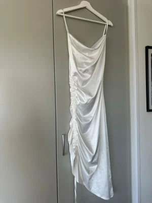 Vit satin klänning med snörning - Snygg vit långklänning i satin med smala axelband och draperad snörning längs sidan. Klänningen har en elegant glansig finish och en figurnära passform som ger en lyxig känsla. Perfekt för dig som vill sticka ut med en stilren look.