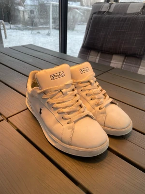Vita Polo Ralph Lauren sneakers - Snygga vita sneakers från Polo Ralph Lauren med klassisk logga på sidan och mörkblå detalj vid hälen. Skorna har lågt skaft, vita snören och är tillverkade i slätt läder. Perfekta för en clean och stilren look. Pris ej hugget i sten!!!