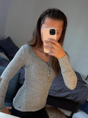 Grå ribbad långärmad tröja Hollister - Säljer en grå ribbad långärmad tröja från Hollister med knappar framtill. Tröjan har en figurnära passform, rund halsringning och är tillverkad i ett mjukt, stretchigt material. Perfekt för lager-på-lager och enkel att matcha med jeans eller kjol.