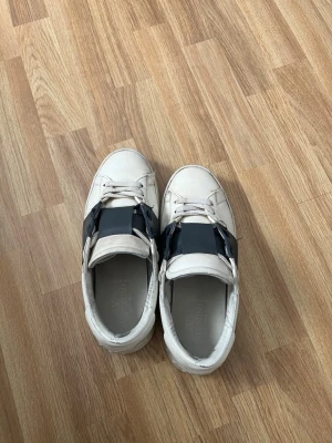 Valentino Garavani vita sneakers - Snygga vita sneakers från Valentino Garavani med en bred svart rem över vristen. Skorna har klassisk låg profil, rund tå och är tillverkade i läder. Kommer med originalkartong och dustbags. Perfekta för dig som vill ha en exklusiv touch på din outfit.