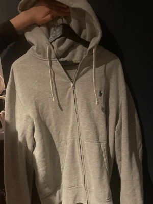 Grå zip hoodie från Polo Ralph Lauren - Snygg gråmelerad hoodie med dragkedja från Polo Ralph Lauren. Klassisk design med huva och vita dragsnören, två fickor framtill och den ikoniska lilla loggan broderad på bröstet. Perfekt för en chill och avslappnad stil.