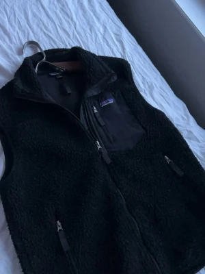 Patagonia väst - Svart fleeceväst från Patagonia med dragkedja framtill, tre fickor med dragkedja och en bröstficka i nylon. Västen har hög krage och är tillverkad i mjukt teddyfleece-material. Perfekt för lager-på-lager och chill stil.