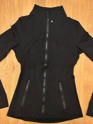 Lululemon Define Jacket - Slimmad svart jacka från Lululemon med hög krage och hel dragkedja framtill. Jackan har två snedställda fickor med dragkedja och långa ärmar. Perfekt för dig som gillar sportig och clean stil. Storlek 2, hellång, luonmaterial. Jag har använt den en gång inomhus. Som ny i skick! Jag har kvittot😊