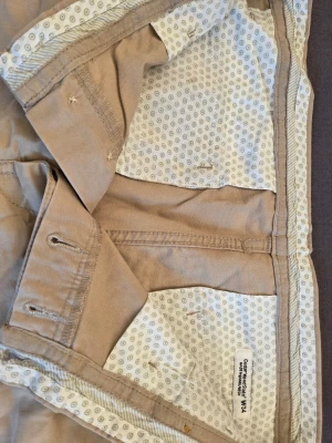 Beige shorts från H&M - Snygga beige shorts från H&M med klassisk passform och midwaist. Tillverkade i mjuk bomull med diskreta fickor och knappgylf. Insidan har ett ljust mönstrat tyg som ger en extra detalj. Perfekta för varma dagar och enkel att matcha.