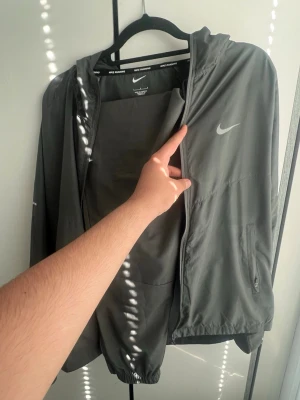 Grå Nike vindjacka med huva - Snygg grå vindjacka från Nike med huva och dragkedja framtill. Jackan har två sidofickor med dragkedja och Nike-logga på bröstet. Perfekt för löpning eller när du vill ha en lätt jacka. Tillverkad i ett tunt, vindtätt material.