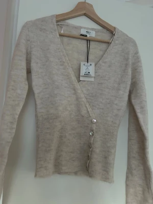 Gina tricot stickad tröja - Säljer en beige, mjuk kofta från Gina Tricot i alpackablandning. Koftan har v-ringning, långa ärmar och sned knäppning med fyra knappar framtill. Perfekt för lager på lager och ger en clean look.