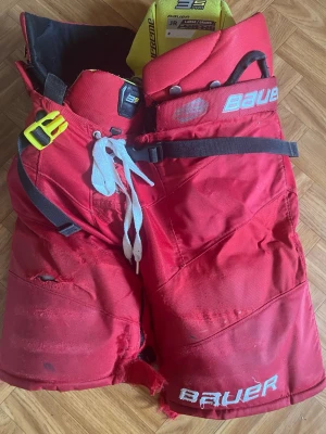 Röda hockeybyxor från Bauer JR - Röda hockeybyxor från Bauer i juniorstorlek Large. Byxorna har snörning i midjan, justerbart bälte och extra vaddering för skydd. Slitstarkt material och klassisk design med Bauer-logga framtill.