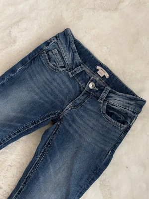 Lågmidjade bootcut jeans - Snygga blå bootcut jeans med lätt slitna detaljer och klassisk femficksdesign. Jeansen har låg midja och är tillverkade i denim med stretch för extra komfort. Perfekta för en avslappnad och trendig look.
