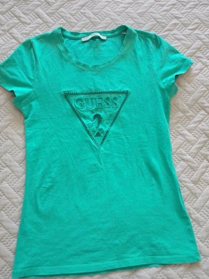 Turkos/Mint t-shirt från Guess - Snygg turkos t-shirt från Guess med klassisk triangel-logga framtill. T-shirten har rund halsringning och korta ärmar. Perfekt för dig som gillar stilrena och trendiga plagg med tydlig logga.
