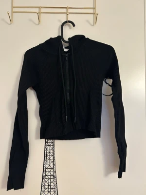 Svart ribbad hoodie med dragkedja från H&M - En svart ribbad hoodie med dragkedja framtill och långa ärmar från H&M i storlek XS. Hoodien har en huva och dragsko samt är croppad i modellen. Materialet är stretchigt och följsamt. 
