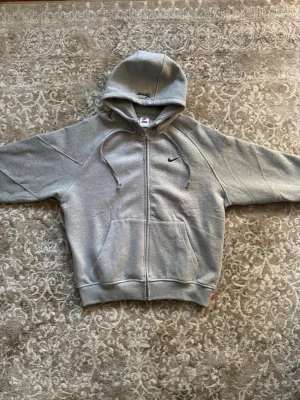 Grå zip hoodie från Nike x Supreme - Snygg grå hoodie med dragkedja från Nike och Supreme. Klassisk huva med broderad Supreme-logga, fickor framtill och liten svart Nike Swoosh på bröstet. Tillverkad i mjuk bomullsblandning, perfekt för en avslappnad streetwear-look.