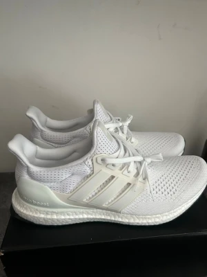Adidas Ultra Boost vita sneakers - Fräscha helvita Adidas Ultra Boost sneakers med stickad ovandel i mesh och klassiska tre ränder på sidan. Skorna har en bekväm Boost-sula och snörning framtill. Perfekta för dig som gillar stilrena och sportiga sneakers.