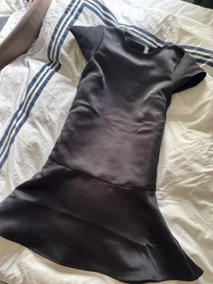 Svart satin klänning från H&M Divided - Supersnygg svart klänning i satin från H&M Divided. Modellen har korta ärmar, rund halsringning och en volang nedtill som ger en extra touch. Perfekt för dig som gillar stilrena och trendiga plagg.