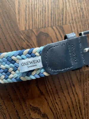 Flätat bälte från ONEWear Sweden - Snyggt flätat bälte från ONEWear Sweden i blå, beige och grå toner. Bältet har detaljer i mörkblått skinn och ett vävt mönster som ger en cool och avslappnad look. Perfekt accessoar för att lyfta din outfit.