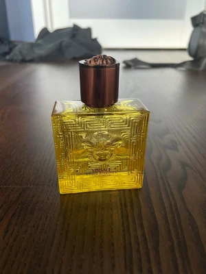 Versace Eros Eau de Parfum - Versace Eros Eau de Parfum i en fyrkantig, transparent flaska med gul vätska och ett elegant mönster. Flaskan har en bronsfärgad, metallisk kork och Medusa-symbolen på framsidan. Perfekt för dig som gillar lyxiga och kraftfulla dofter.