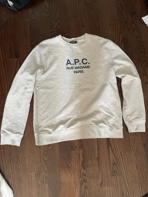 Grå sweatshirt från A.P.C. - Säljer en stilren grå sweatshirt från A.P.C. med tryck i mörkblått på bröstet där det står 'A.P.C. RUE MADAME PARIS'. Tröjan har rund halsringning, långa ärmar och ribbade muddar. Perfekt för en clean och avslappnad look.